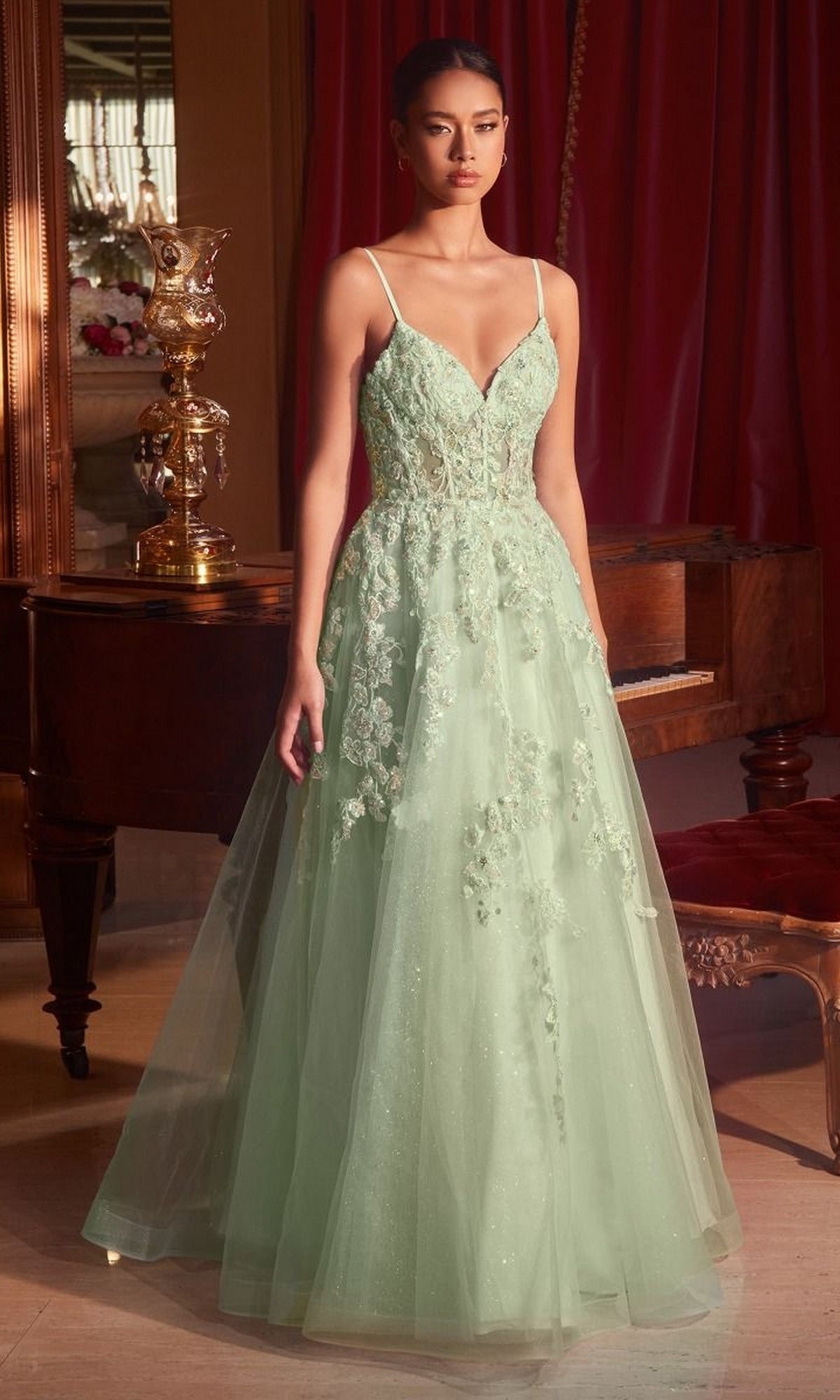 A woman stands indoors in the Long Prom Ball Gown with Flowers CM347, a light green floor-length dress with floral embellishments and a tulle skirt. A grand piano and red curtains complete the elegant scene.